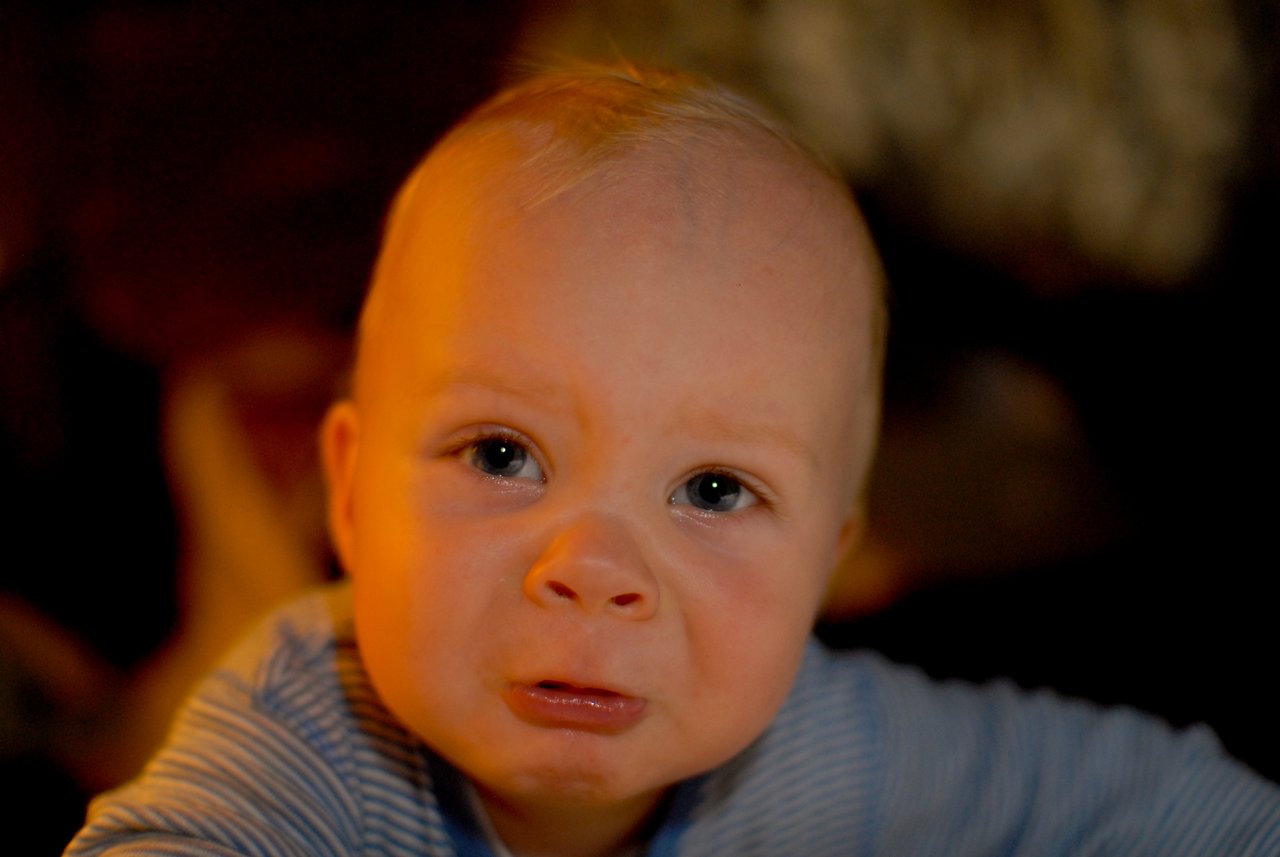 A baby with a slightly distressed expression looks directly at the camera, wearing a striped outfit in dim lighting.