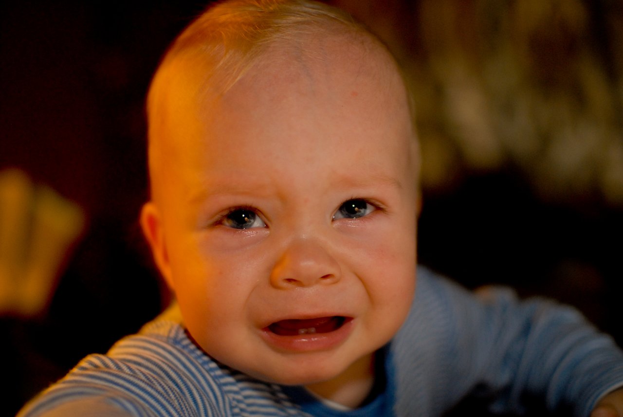A baby with light skin and short blond hair is crying, wearing a blue striped outfit indoors.