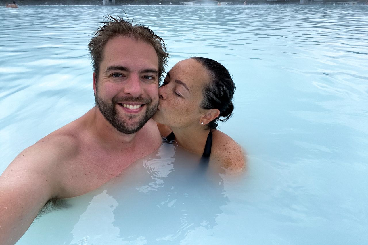 A couple in the Blue Lagoon; one person smiles at the camera while the other kisses their cheek.