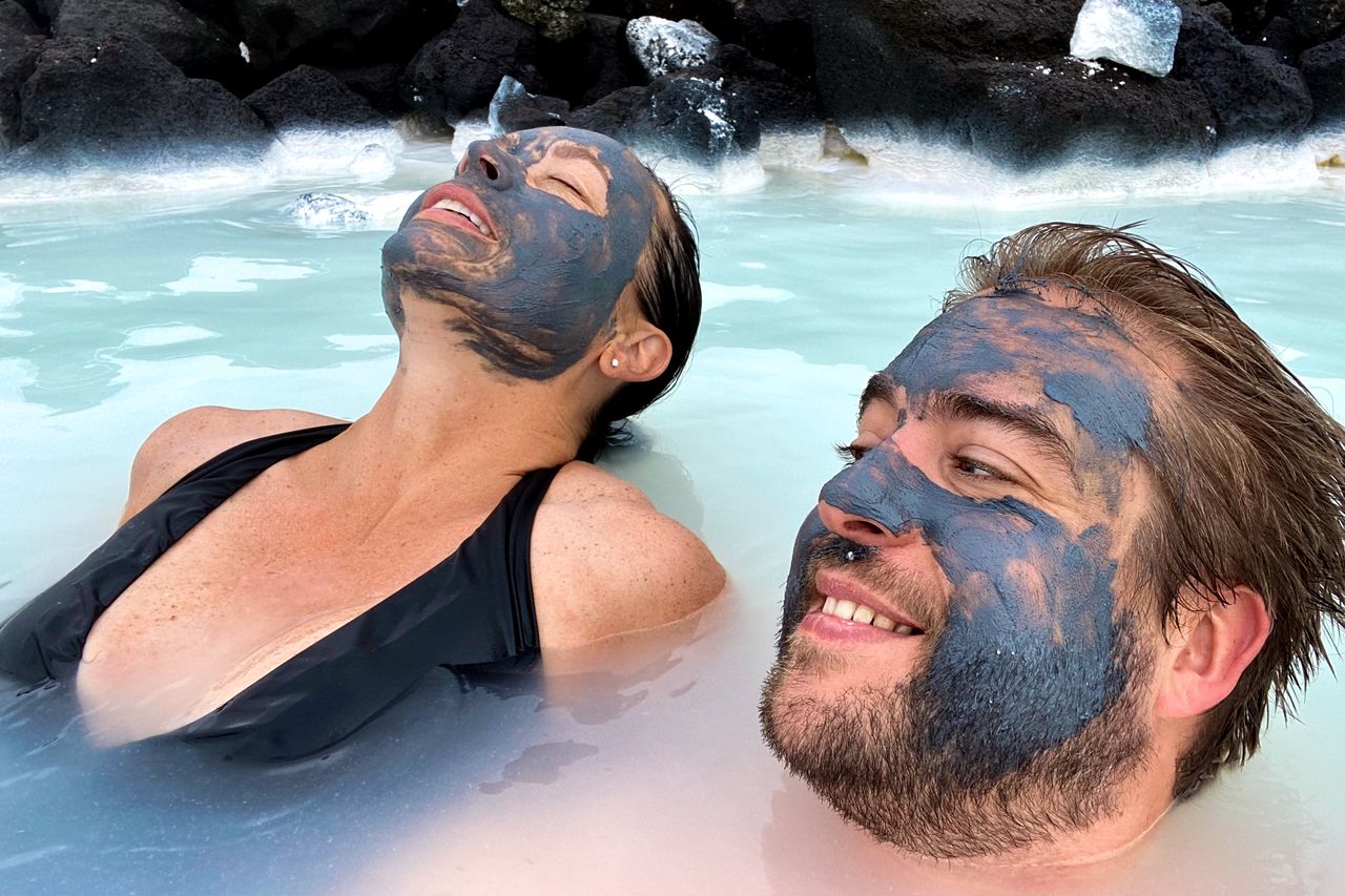 Two people relax in the Blue Lagoon, Iceland, with dark facial masks while floating in the milky blue water.