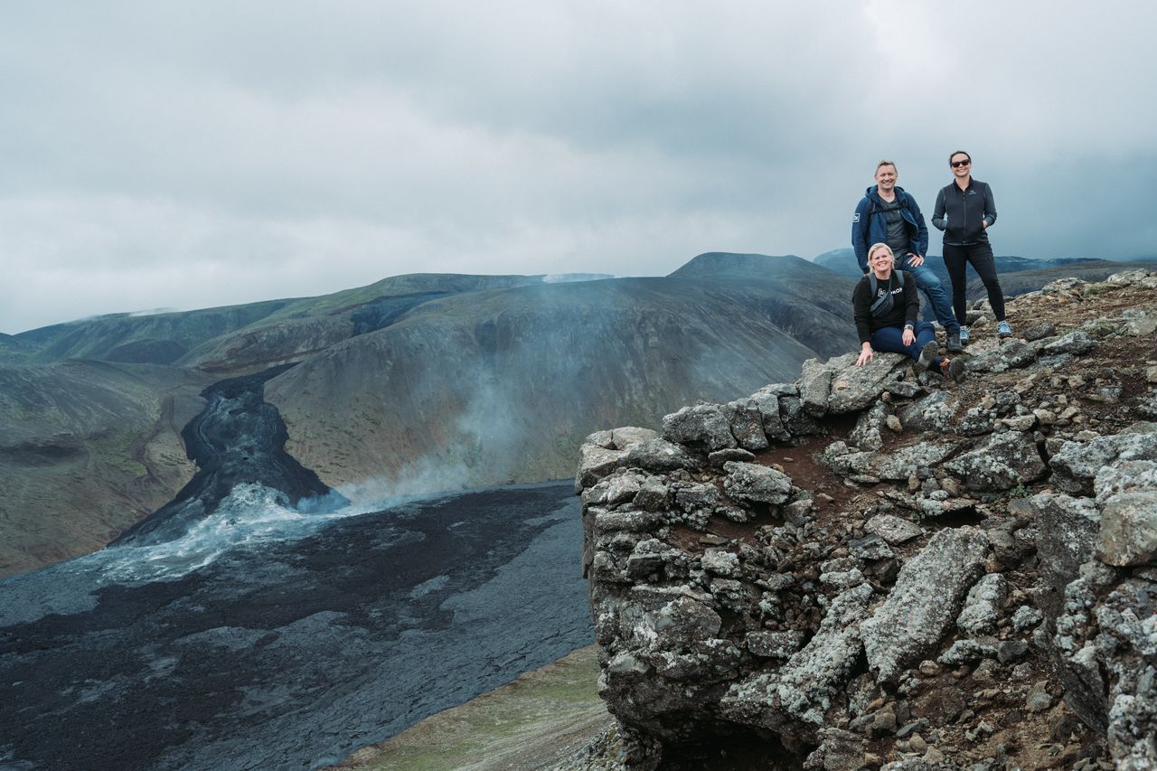 Three people pose on a rocky hill with the smoking Gerlingadalur volcano and lava flow in the background.