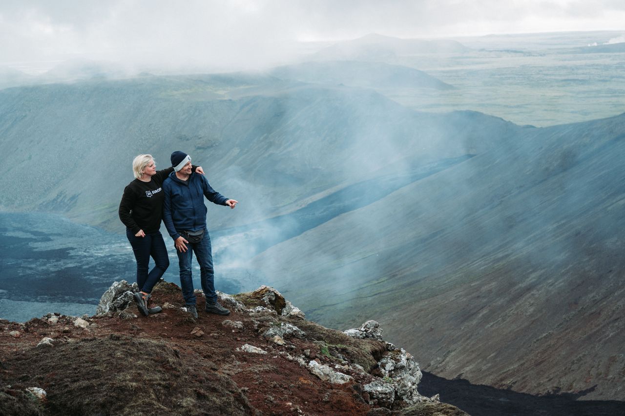 Two people stand on a rocky hill, overlooking the steaming Gerlingadalur volcano in Iceland.