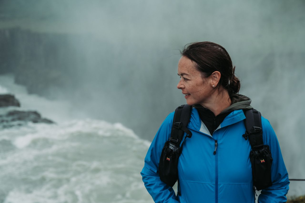 A person wearing a blue jacket and backpack stands near Gullfoss Falls, looking to the side.