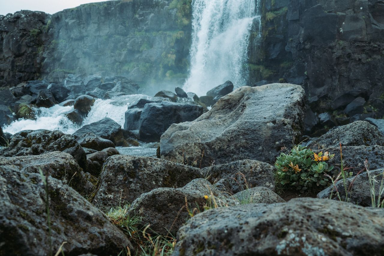 A waterfall cascades down a rocky cliff, with water flowing over large boulders in the foreground.