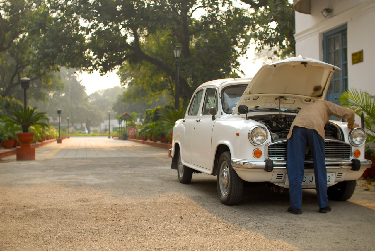 A man inspects the engine of a white Ambassador car with its hood open on a paved driveway.