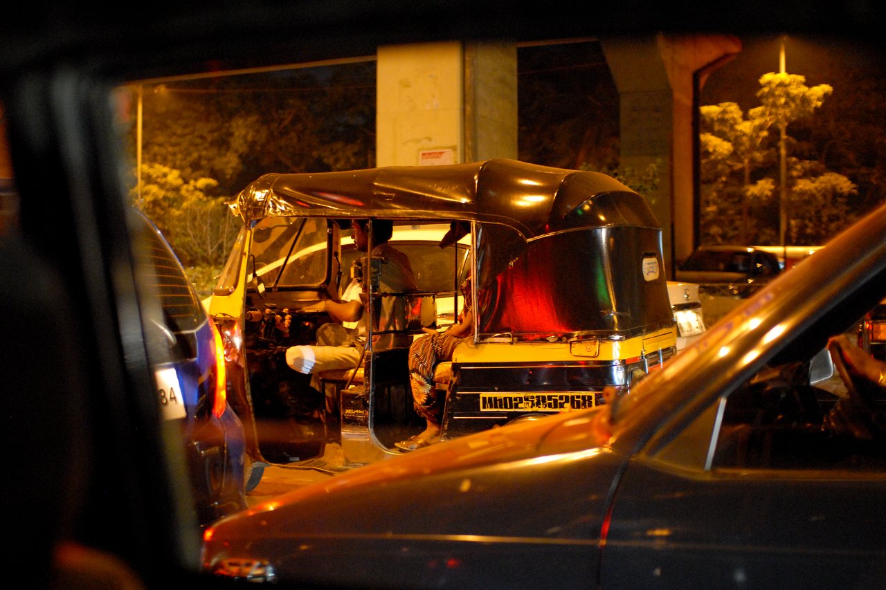 A black and yellow auto rickshaw is stopped in traffic at night, with the driver sitting inside.