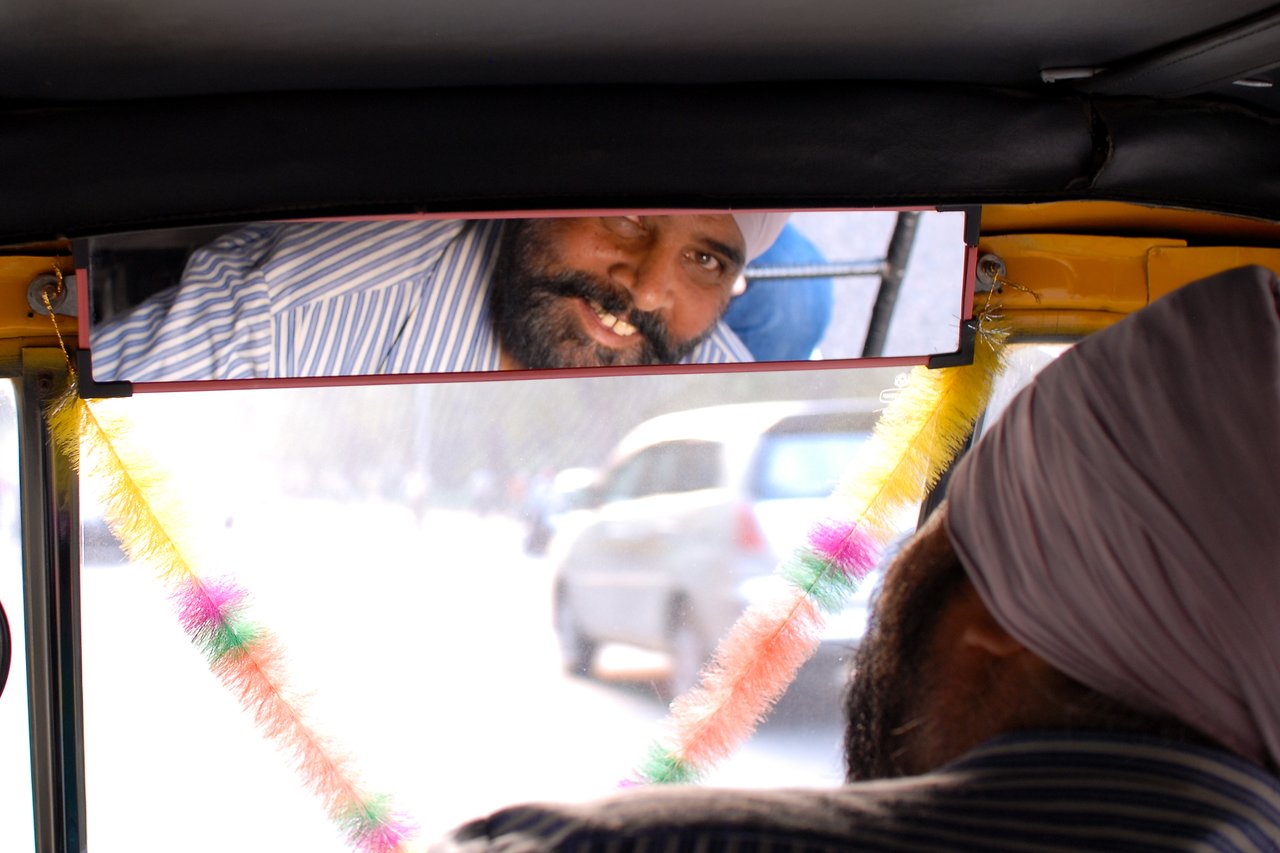 A bearded auto rickshaw driver wearing a turban is seen smiling in the rearview mirror while driving.