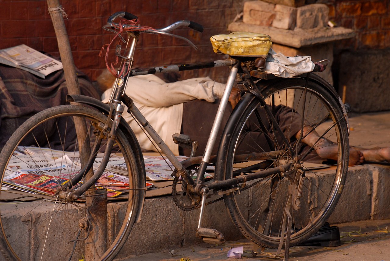 A black bicycle with a basket is parked on the sidewalk, with a person resting on the ground behind it.