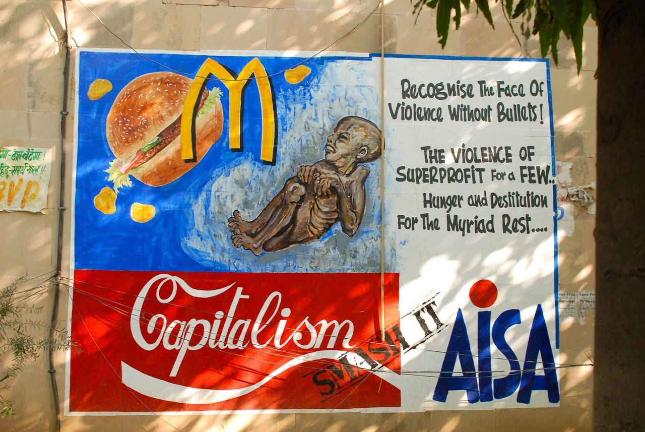 A mural criticizes capitalism, depicting a starving child, a fast-food logo, and text about profit-driven violence and inequality.