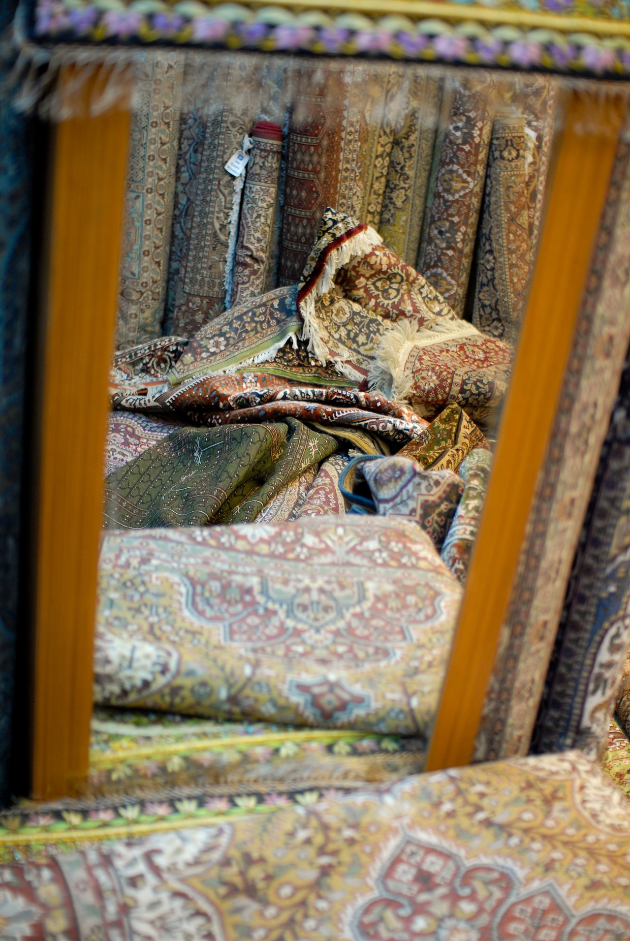 A mirror reflects a display of patterned carpets, with several rolled-up and folded rugs stacked in the background.