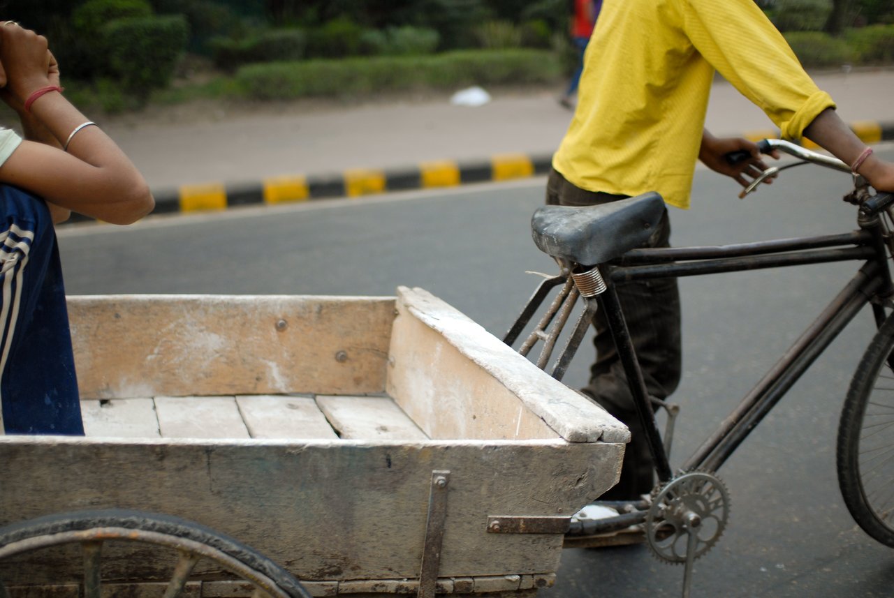 A person pedals a bicycle pulling a wooden cart, while another person sits inside with their arm resting.
