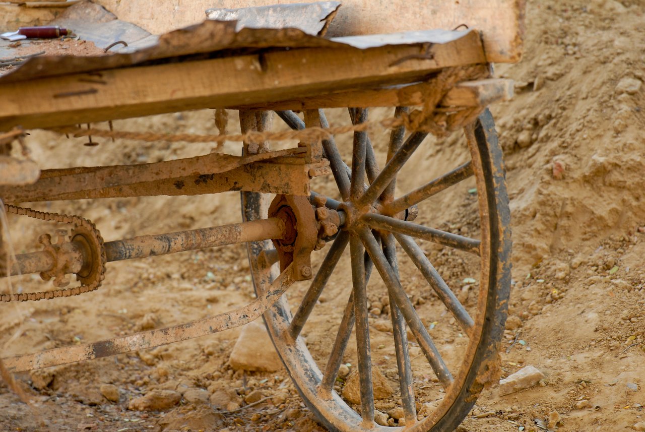 A close-up of an old wooden cart with a metal wheel, covered in dirt and rust.