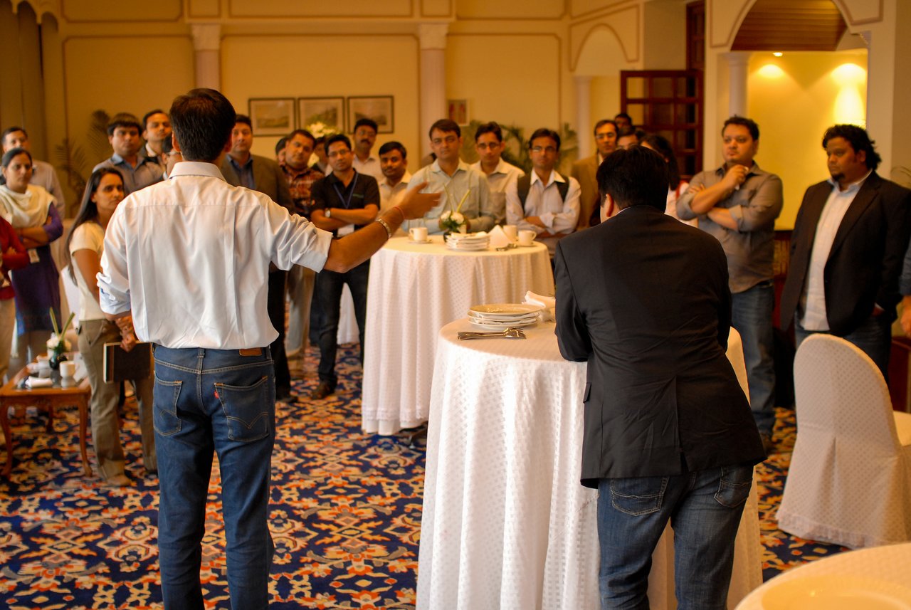 A man in a white shirt gestures while speaking to a group of people gathered around tables in a conference room.