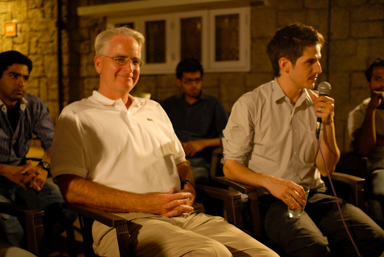 A man in a white shirt smiles while another man speaks into a microphone during a discussion.