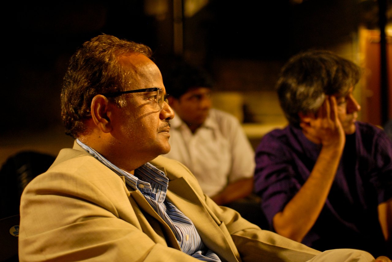 A man in a beige suit listens attentively during a discussion, with two others seated in the background.