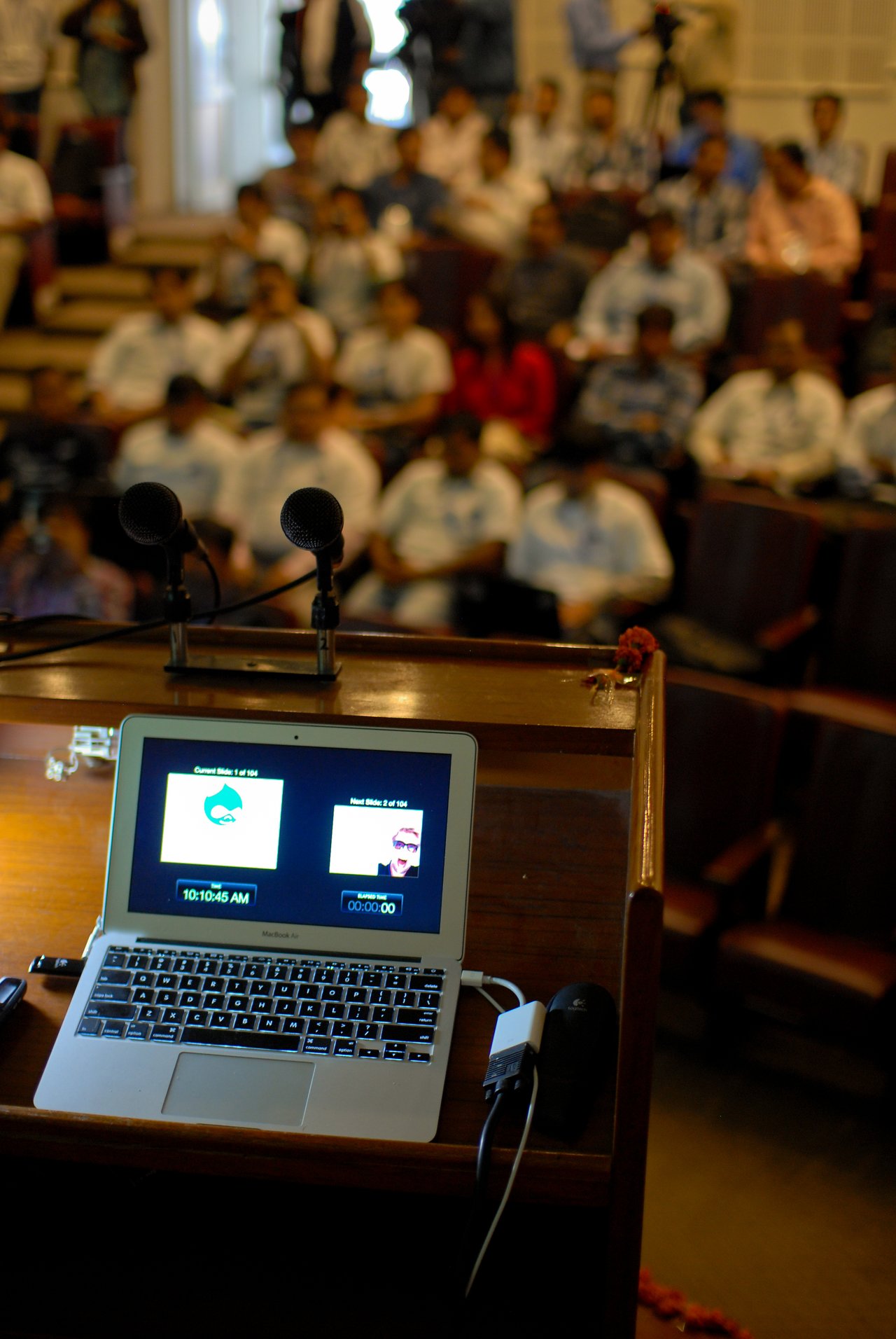 A laptop on a podium displays presentation slides for a keynote at Drupalcamp Delhi, with an audience seated.