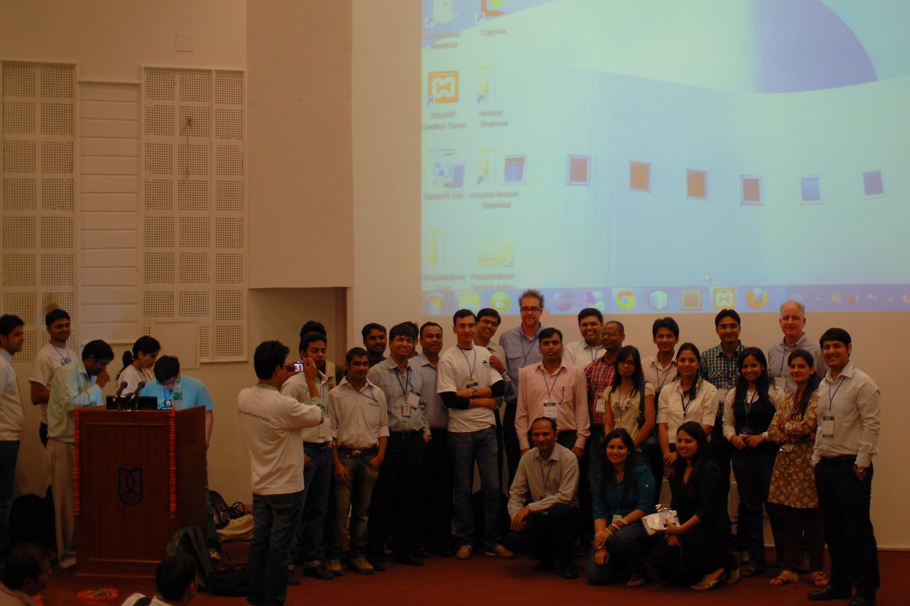 A group of Drupalcamp Delhi organizers poses for a photo on stage, with a large screen in the background.