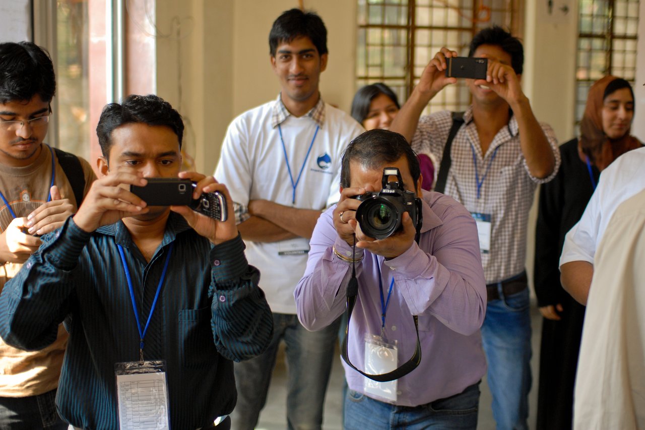 A group of people with cameras and smartphones take photos at a DrupalCamp Delhi event.