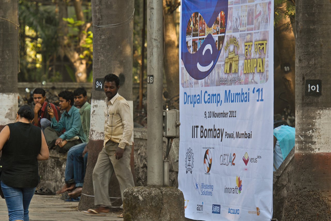 A large banner for Drupal Camp Mumbai 2011 is displayed outdoors, showing event details and sponsor logos.