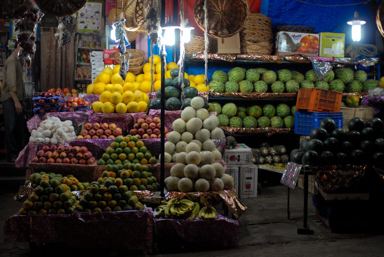 A fruit store with neatly stacked piles of melons, apples, oranges, and watermelons under bright lights at night.