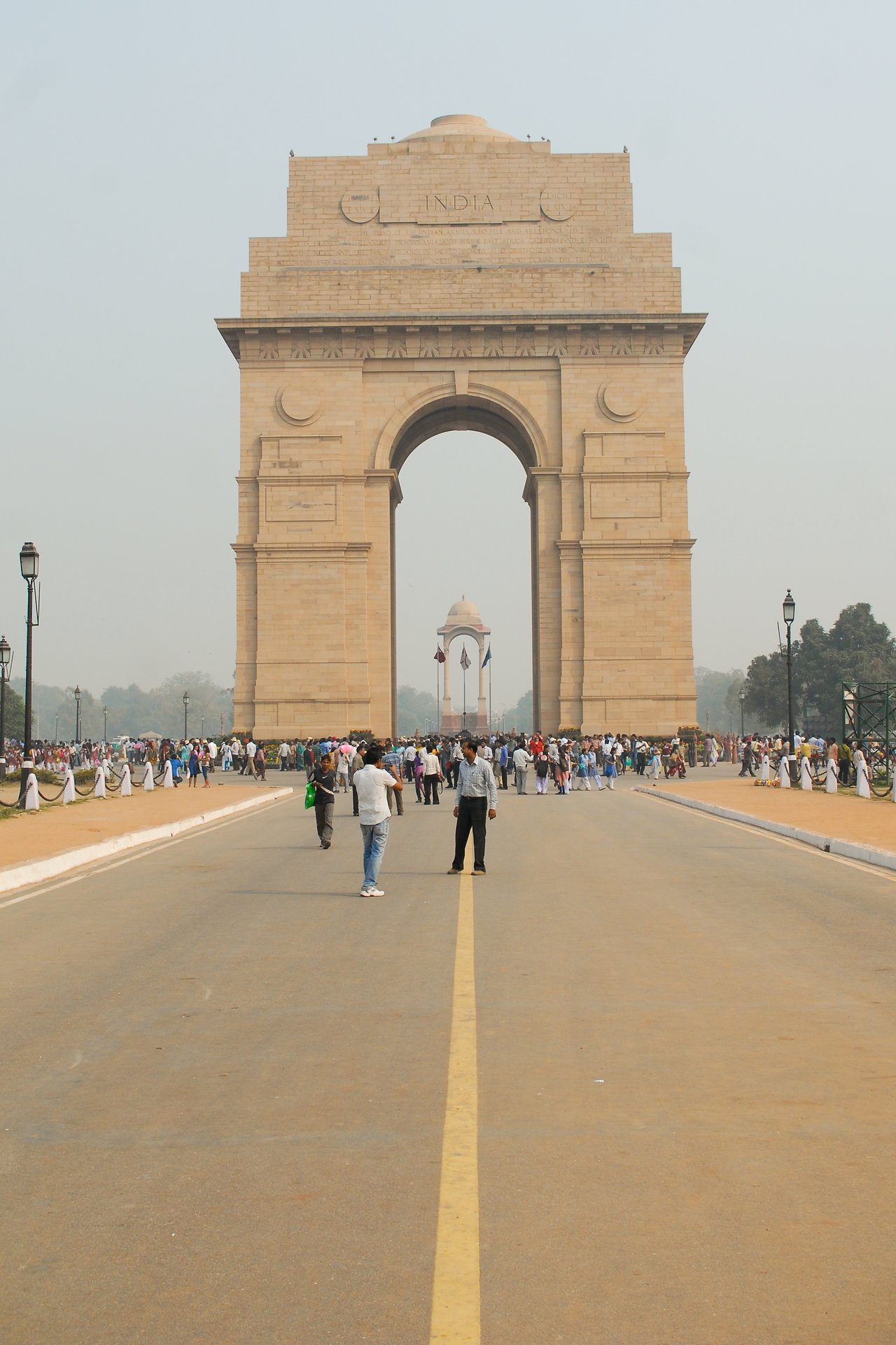 India Gate in New Delhi with many visitors walking around and taking photos on a wide road leading to it.