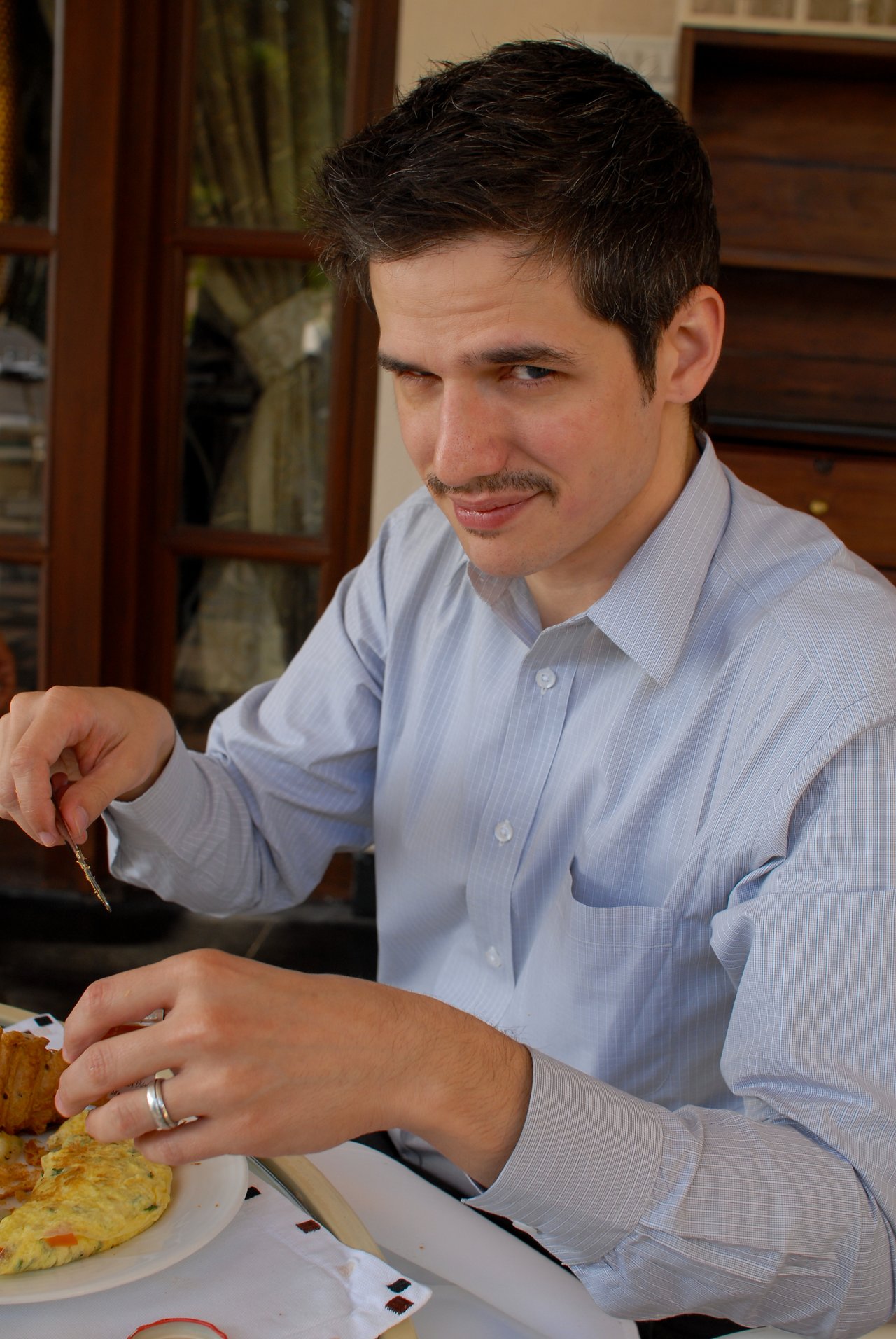 A man in a light blue shirt eats a meal while giving a playful, raised-eyebrow expression.