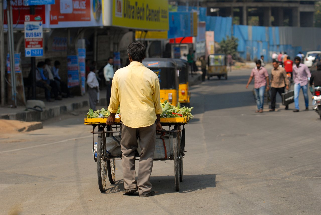 A man in a yellow shirt pushes a cart loaded with limes through a busy street.