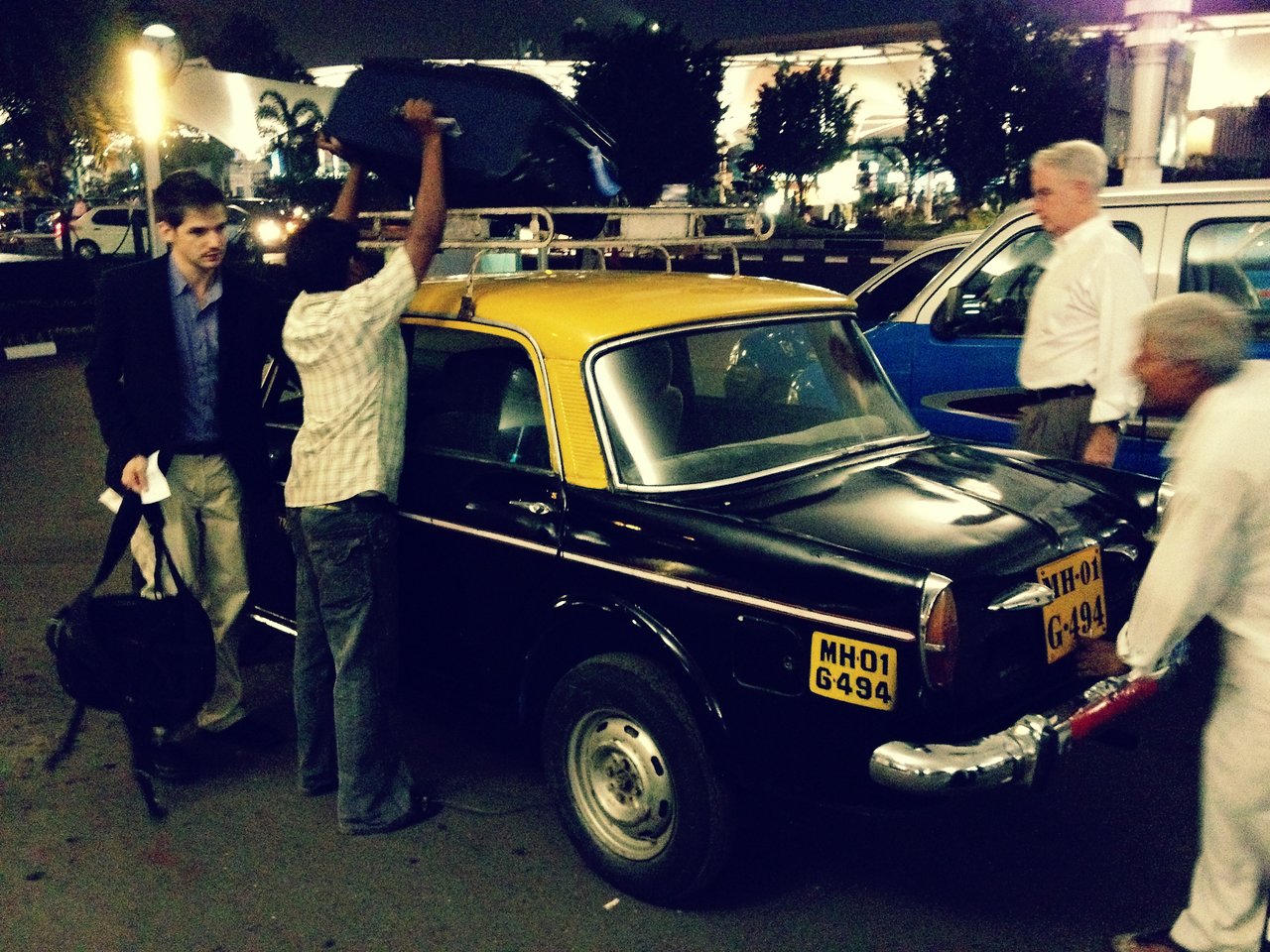 A man lifts a suitcase onto the roof of a black and yellow taxi while others stand nearby.