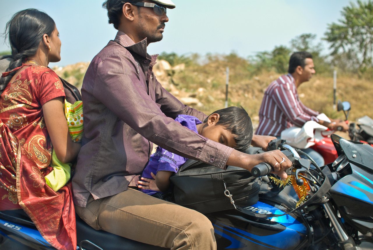 A young child sleeps on the front of a moving motorcycle, resting their head on a bag.