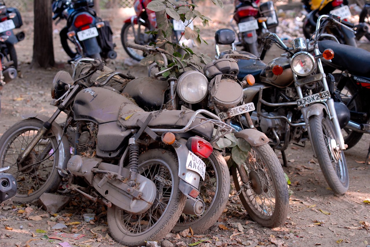 Several dusty, abandoned motorcycles are parked closely together in an outdoor lot, with leaves and debris covering them.