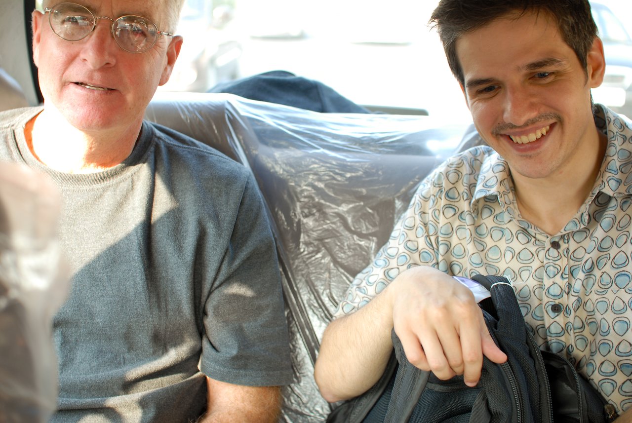 Two men sitting in a car, one wearing glasses and a gray shirt, the other smiling and holding a bag.