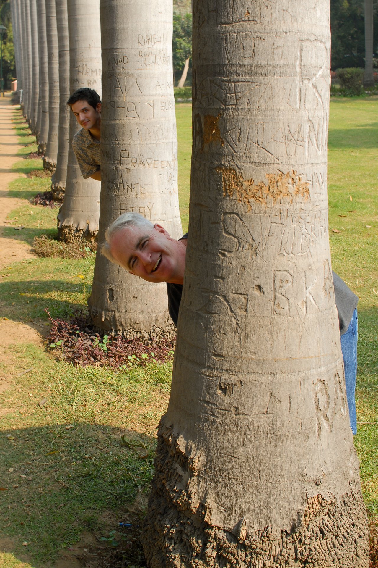 Two people playfully peek out from behind carved tree trunks in a park, smiling at the camera.