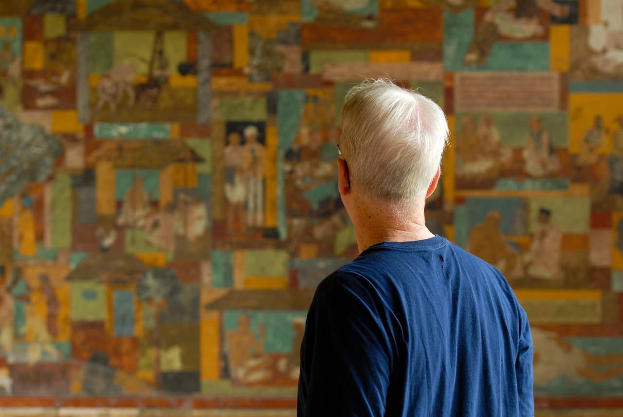 A man with white hair and a blue shirt looks at a colorful mural depicting historical scenes.