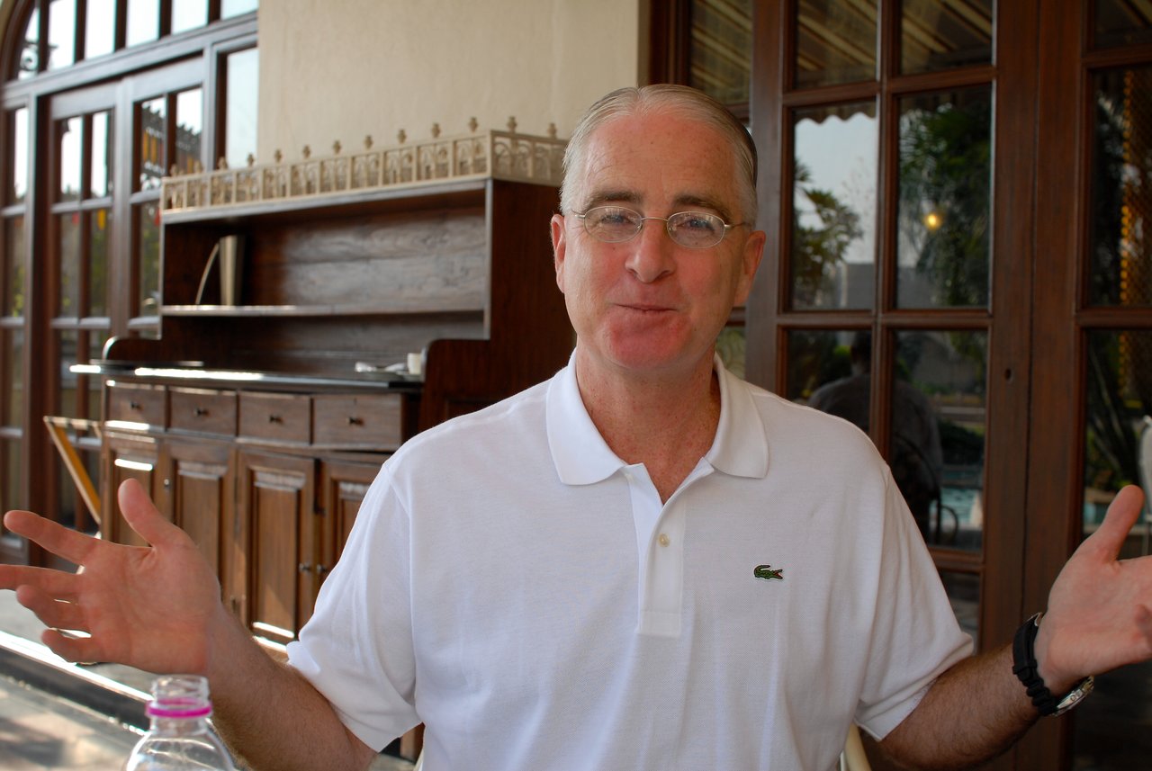 A man in a white polo shirt sits at a table, gesturing with both hands and smiling.