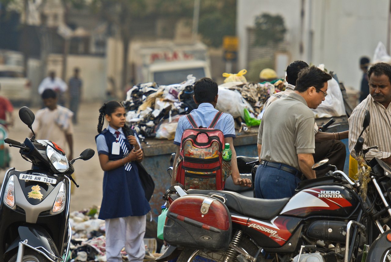 A schoolgirl in a blue uniform stands near a pile of garbage, holding her bag and looking to the side.