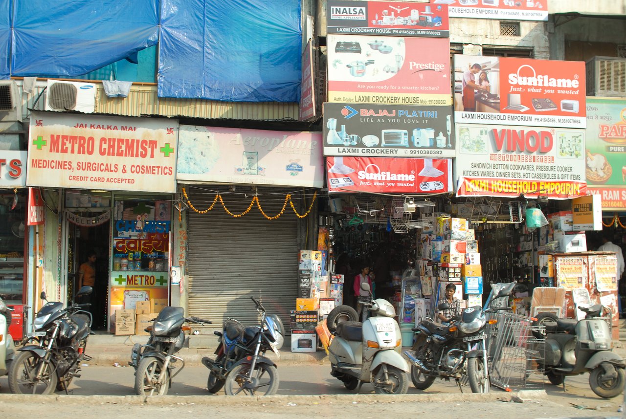 A row of small shops with signs selling medicine, cookware, and household items, with parked motorcycles and scooters in front.