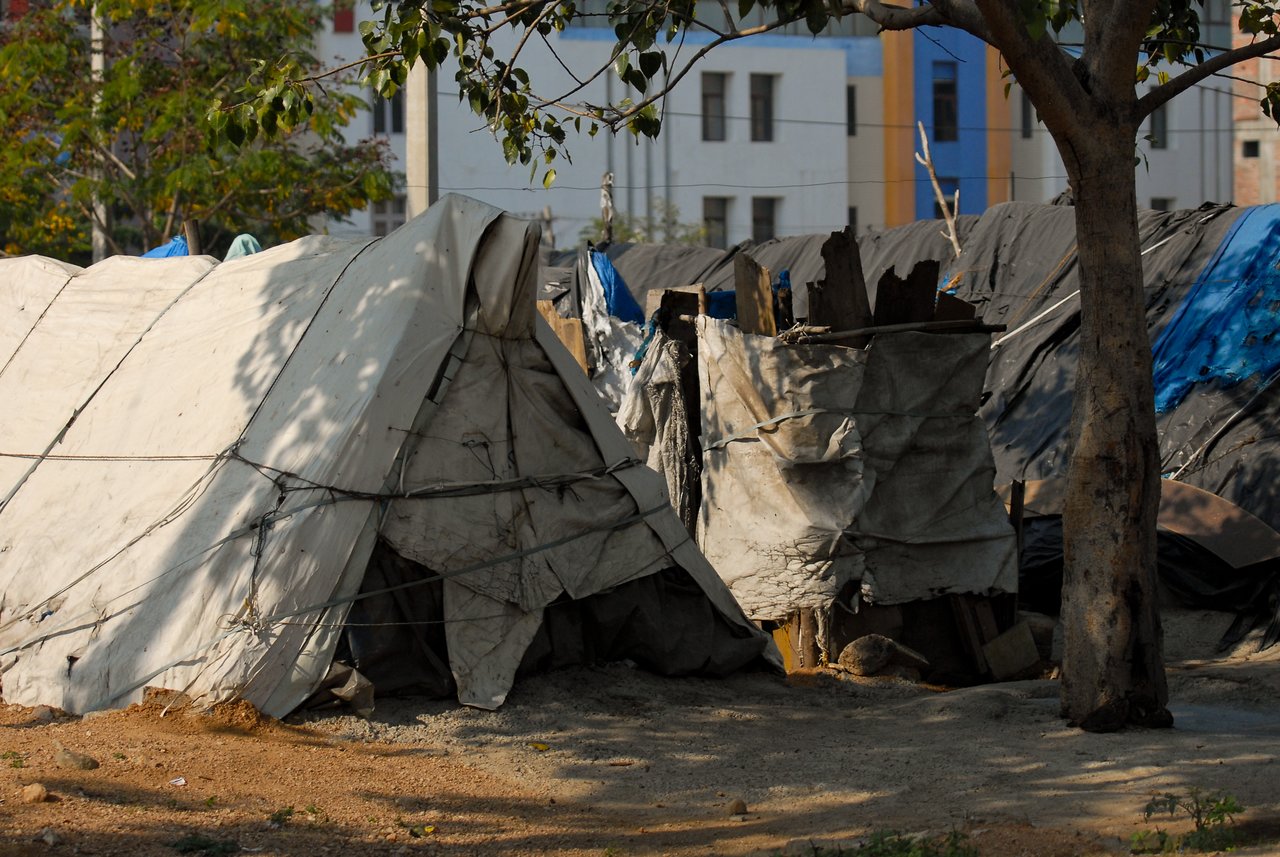 Makeshift tents and shelters made of tarps and cloth in a slum area, with buildings in the background.