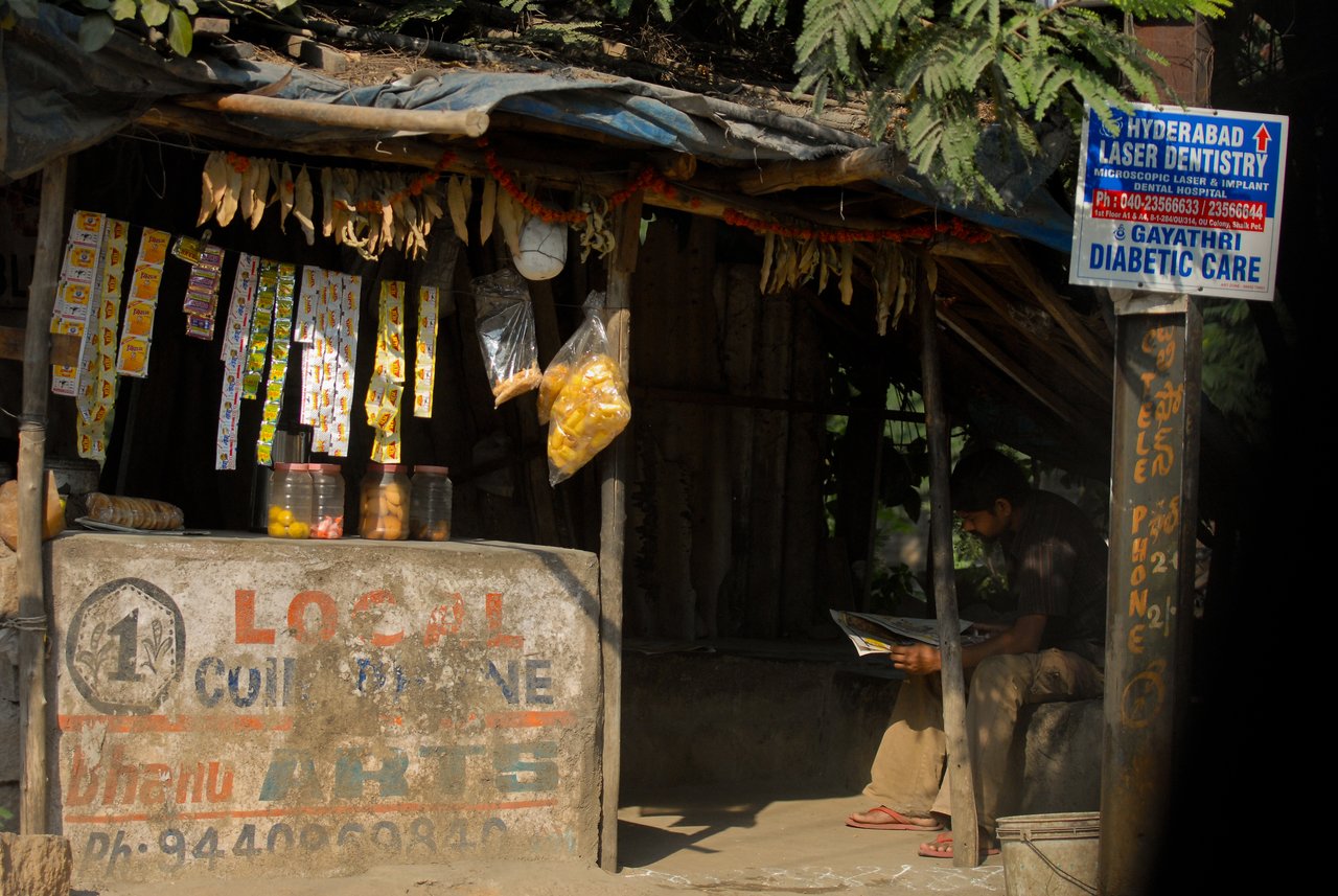 A small roadside shop selling snacks and essentials, with a man sitting nearby reading a newspaper.