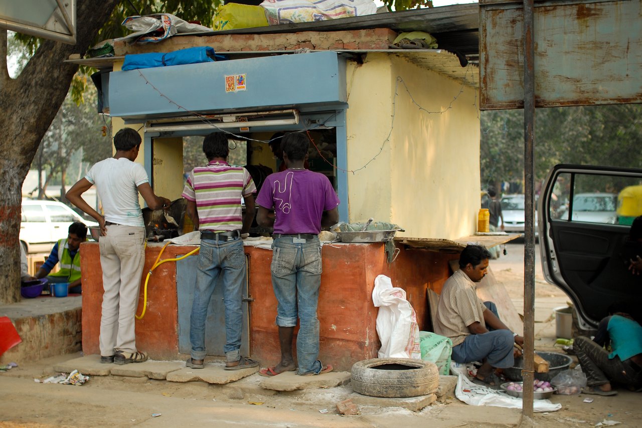 People stand at a small street food stall, ordering and eating, while others sit nearby.