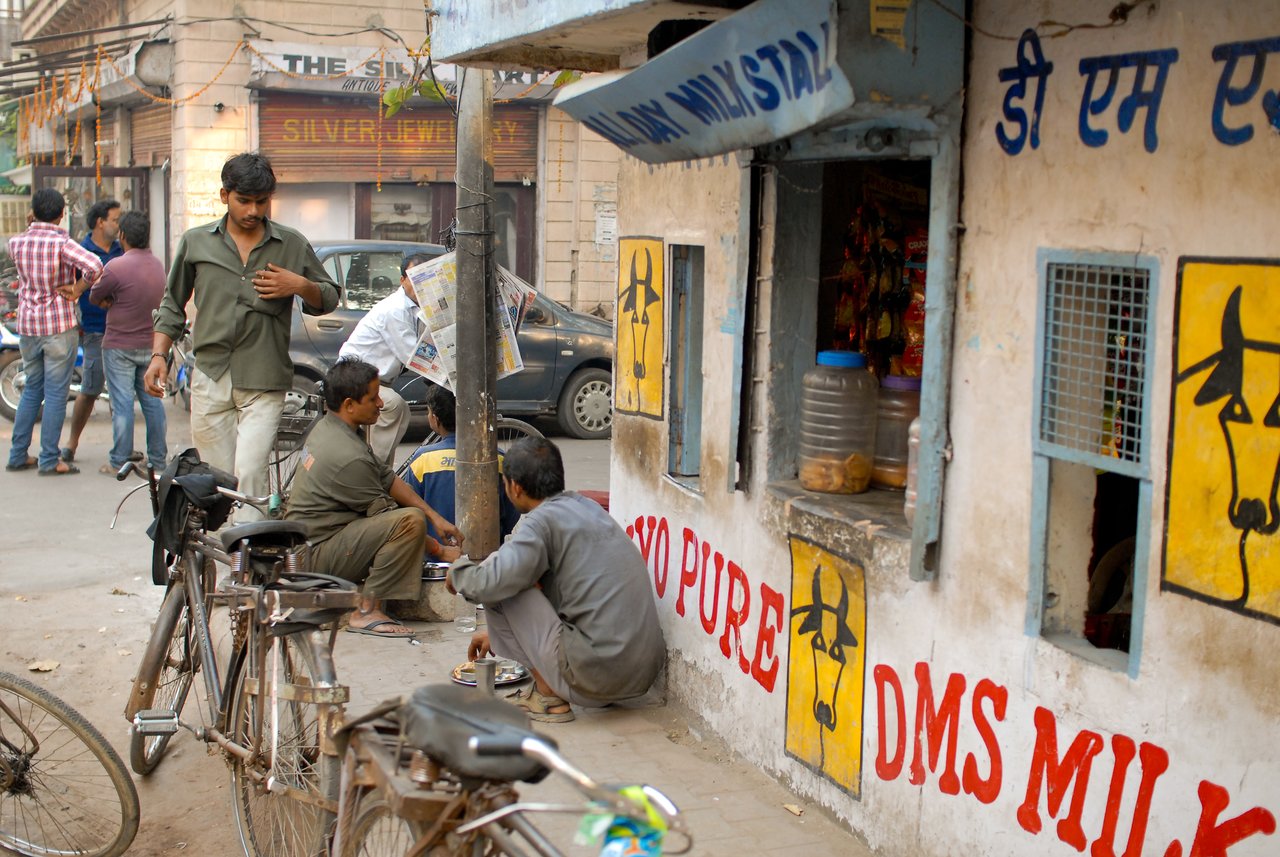 Men sit and eat near a small street food stall with painted signs, while others stand and walk nearby.