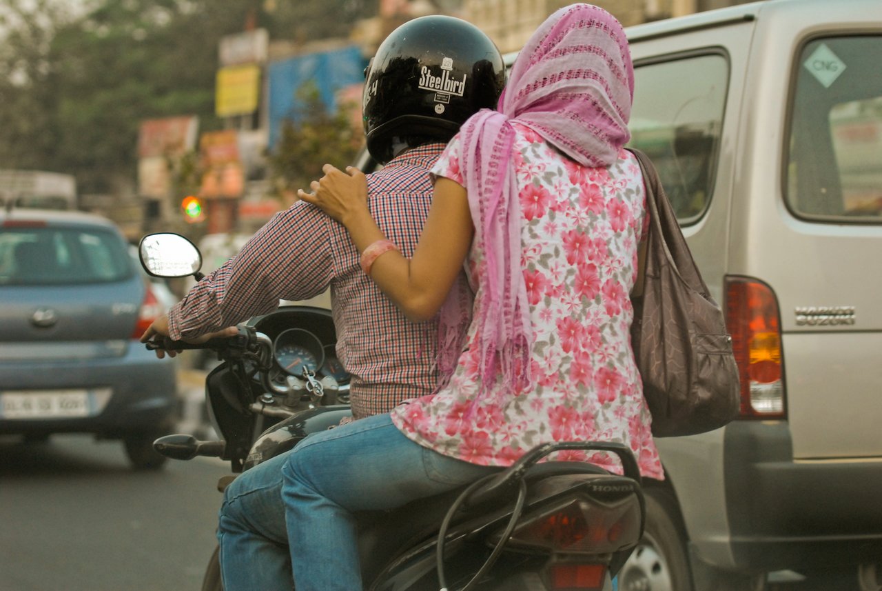 A man wearing a helmet rides a motorcycle with a woman sitting behind him in busy traffic.