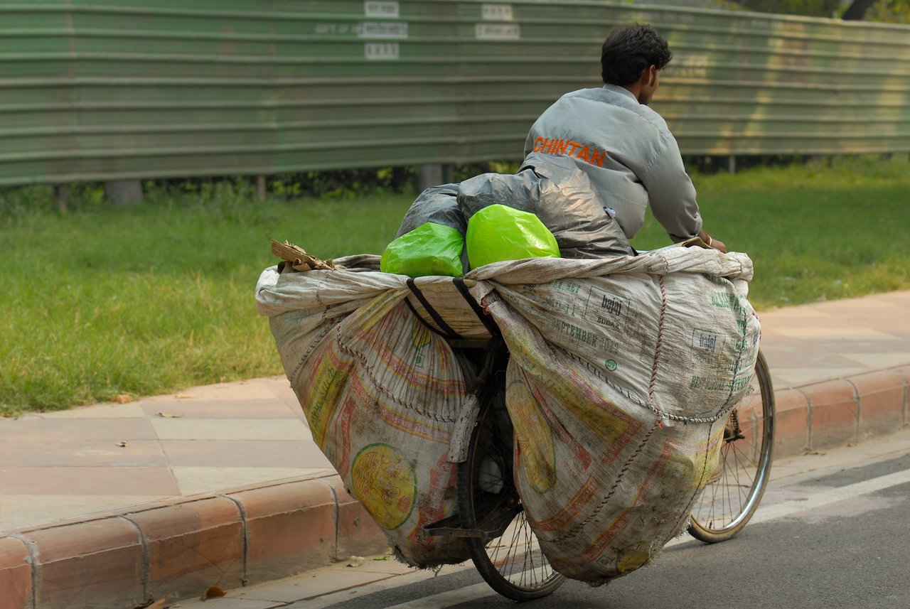 A man rides a bicycle loaded with large sacks and bags, likely transporting goods or recyclables on a city road.