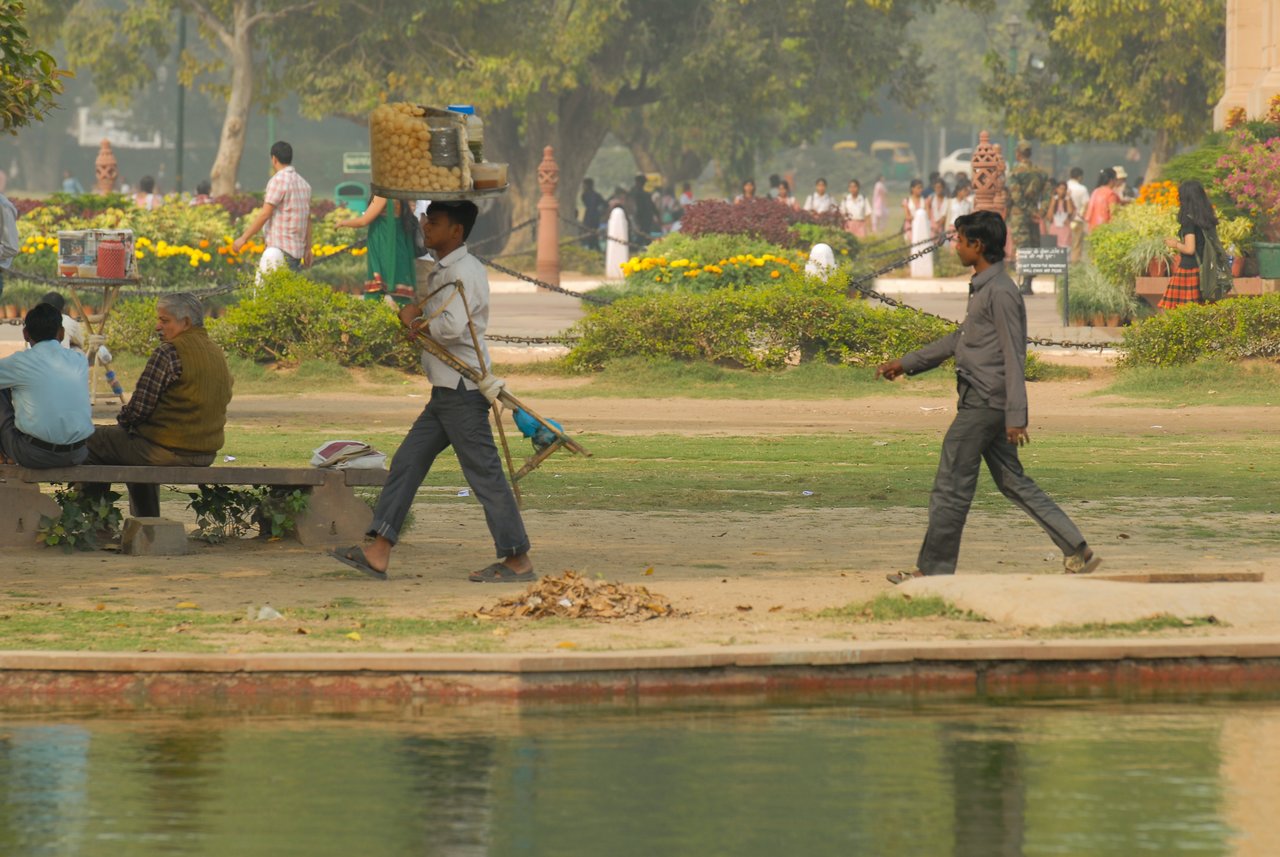 Two men walking in a park, one carrying a basket of snacks on his head and holding a stand.
