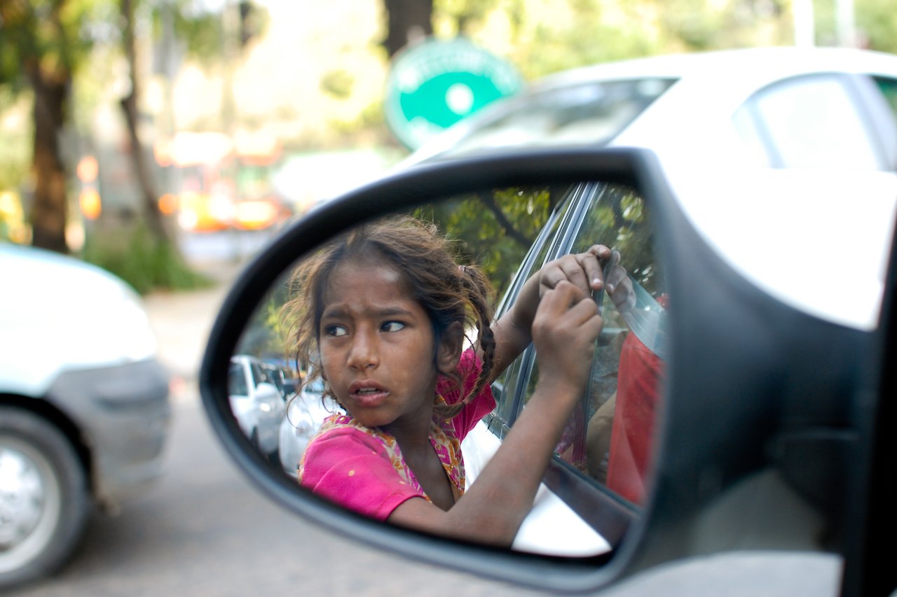 A young girl in a pink dress knocks on a car window, asking for help.