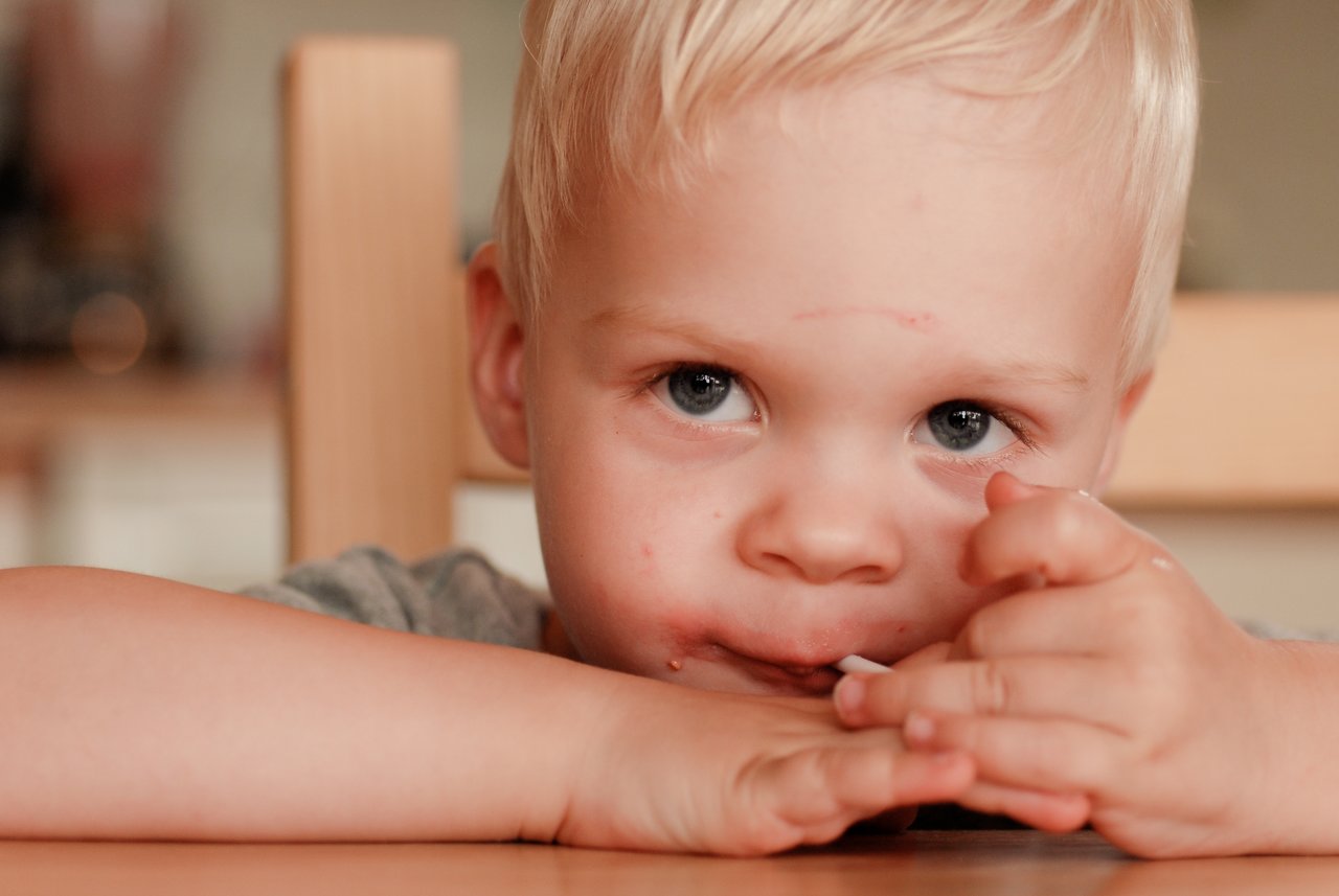 A young child with blonde hair sits at a table, resting their arms and looking directly at the camera.