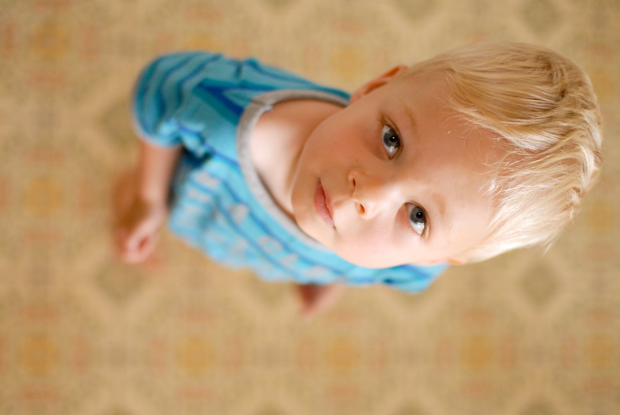 A young child with blonde hair and a blue striped shirt looks up toward the camera with a curious expression.