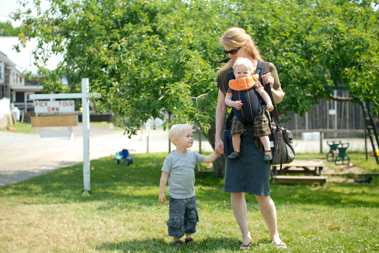 A woman walks outside holding a young child's hand while carrying a baby in a front carrier.