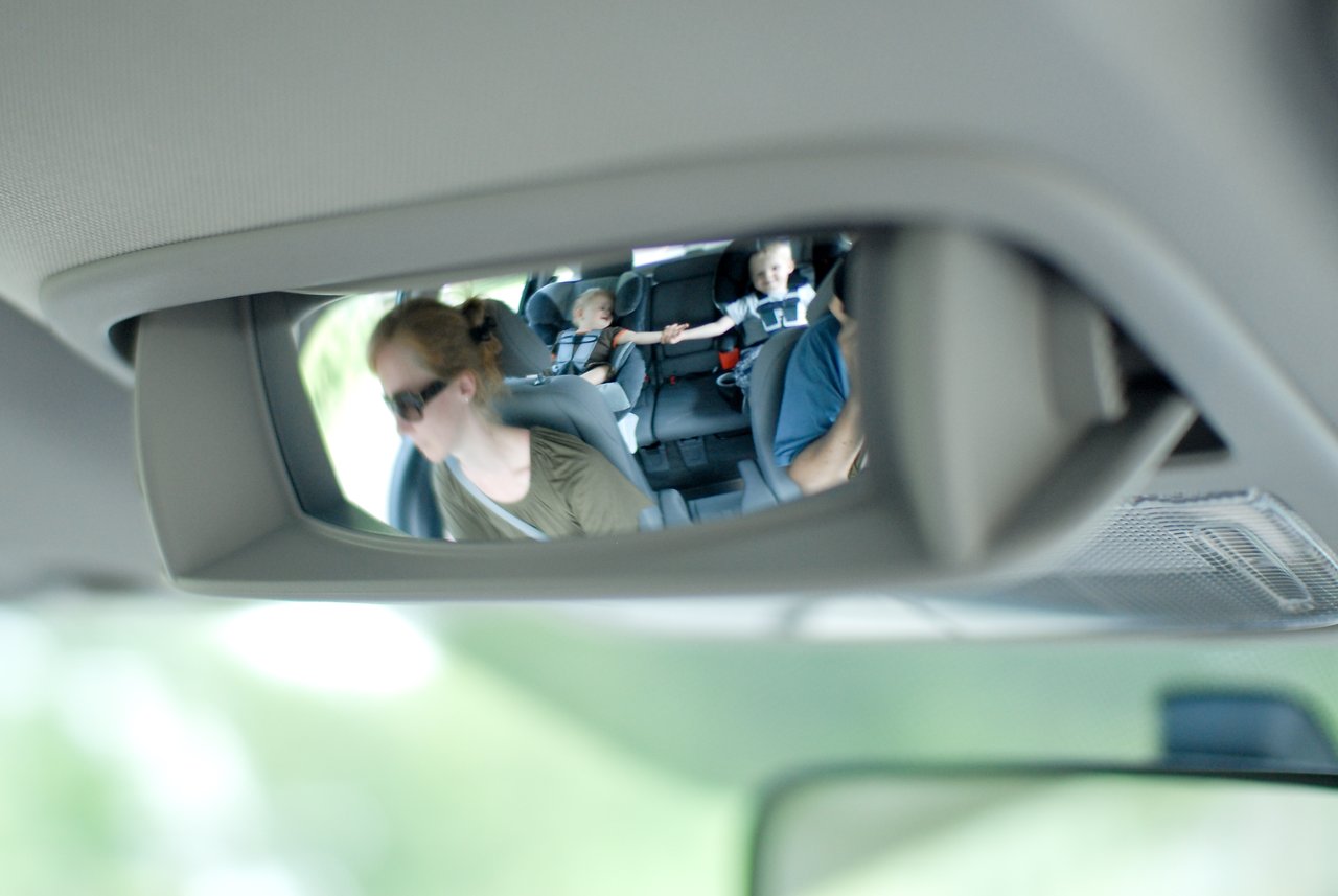 A car's rearview mirror reflects a child in a car seat reaching out, with adults in the front seats.