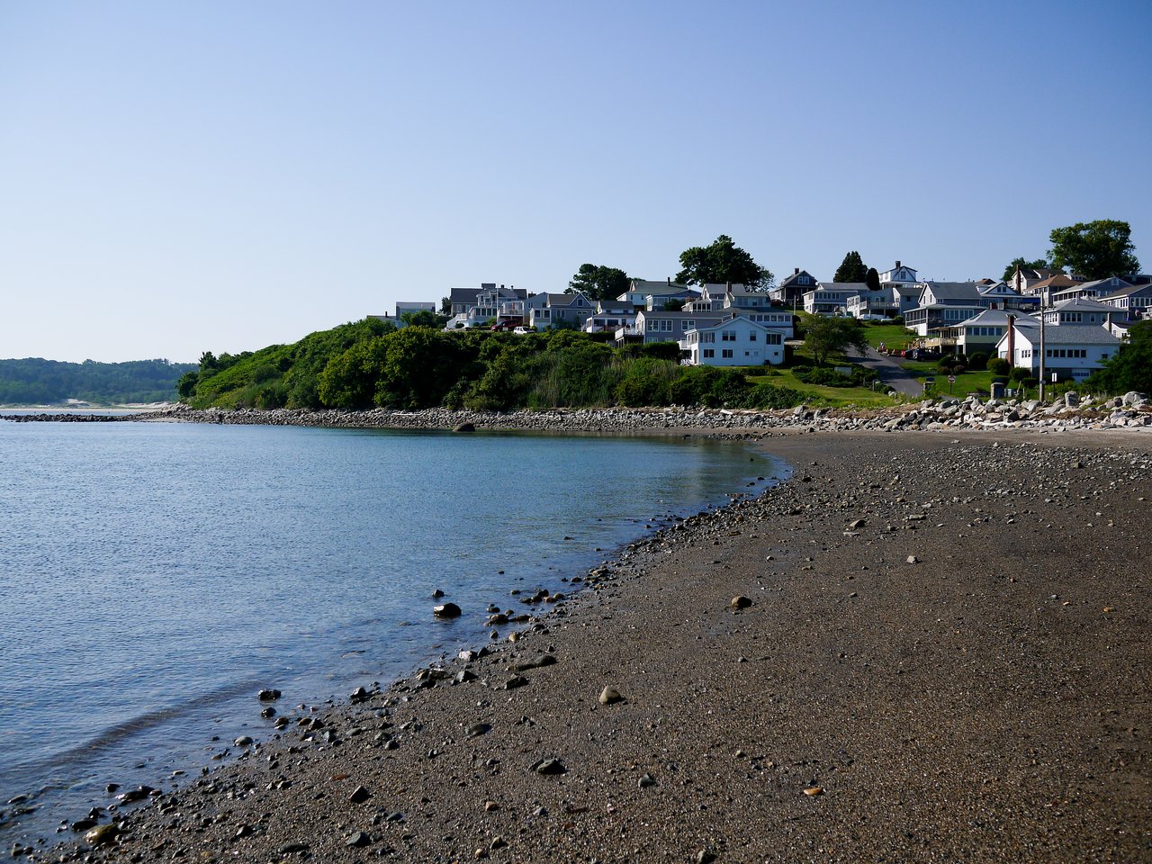 A rocky beach with calm water, with houses on a hill in the background under a clear sky.