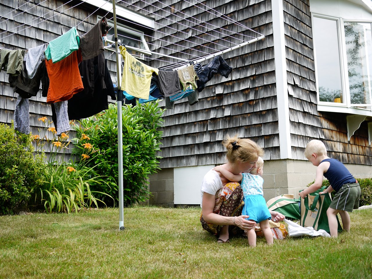 A woman crouches outside with two young children near a laundry bag, while clothes hang on a drying line.